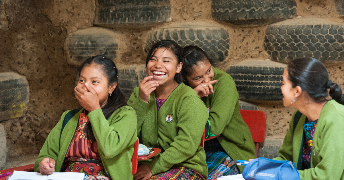 四名身穿绿色毛衣的女学生在用轮胎和泥巴搭成的教室里开怀大笑。
