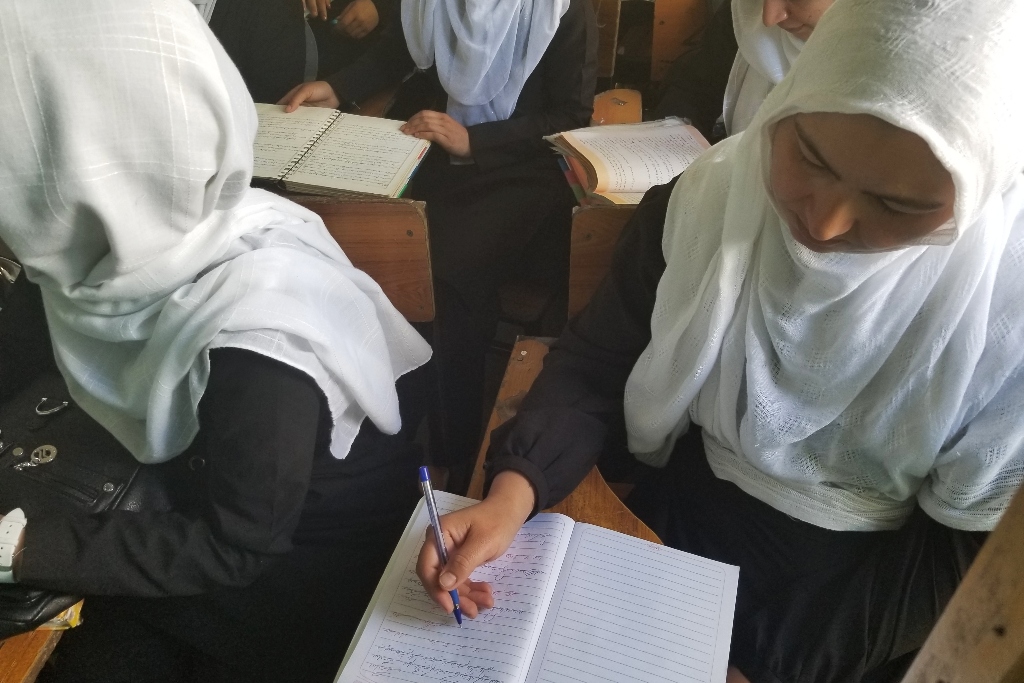 一个戴着白色头巾的女孩在学校课桌前的笔记本上写字。其他学生在她周围。难民的故事