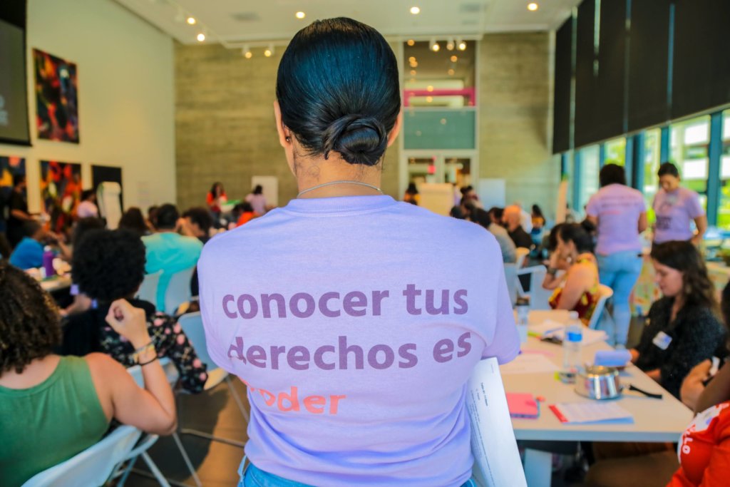 一个穿着紫色t恤的女人站在坐在桌子前的人们前面，t恤后面写着“conocer tus derechos es poder”。
