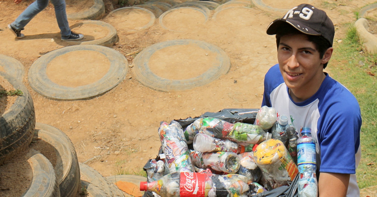 一个小男孩拿着一袋罐头和瓶子靠在轮胎墙上，这是一个创新教育项目的一部分，教孩子们把垃圾变成社区基础设施