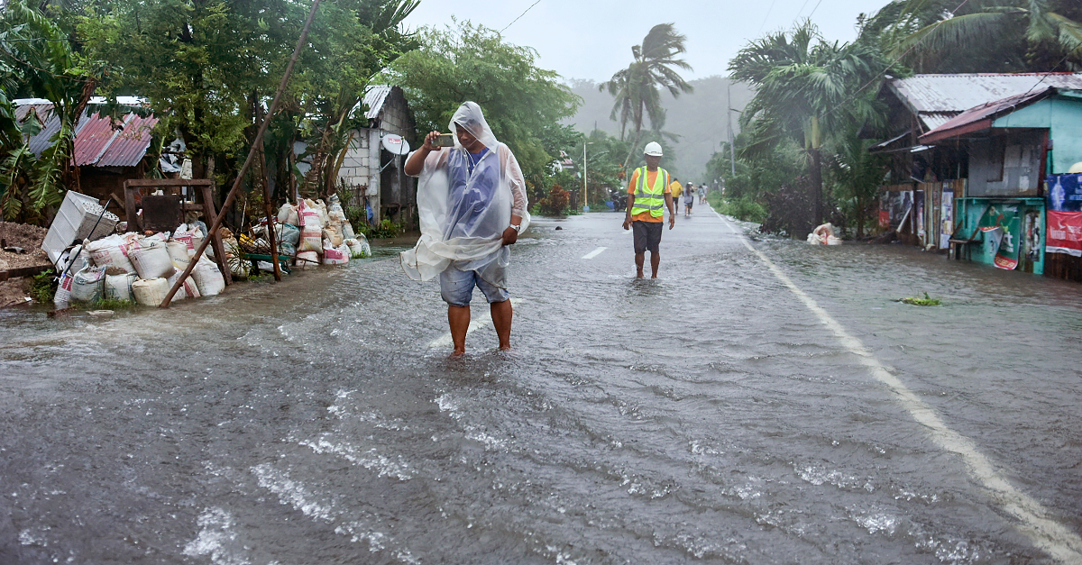 一个穿着透明雨披的人站在被洪水淹没的街道中间，拿着手机拍摄这一场景。一名穿着安全背心的救援人员站在他们身后的路上。