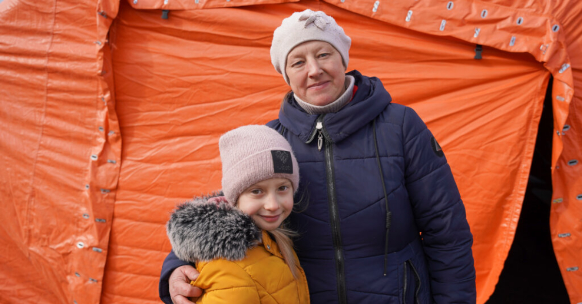 一名妇女和她的女儿在橙色帐篷前穿着冬季夹克和帽子。母亲的手臂搂着女儿。