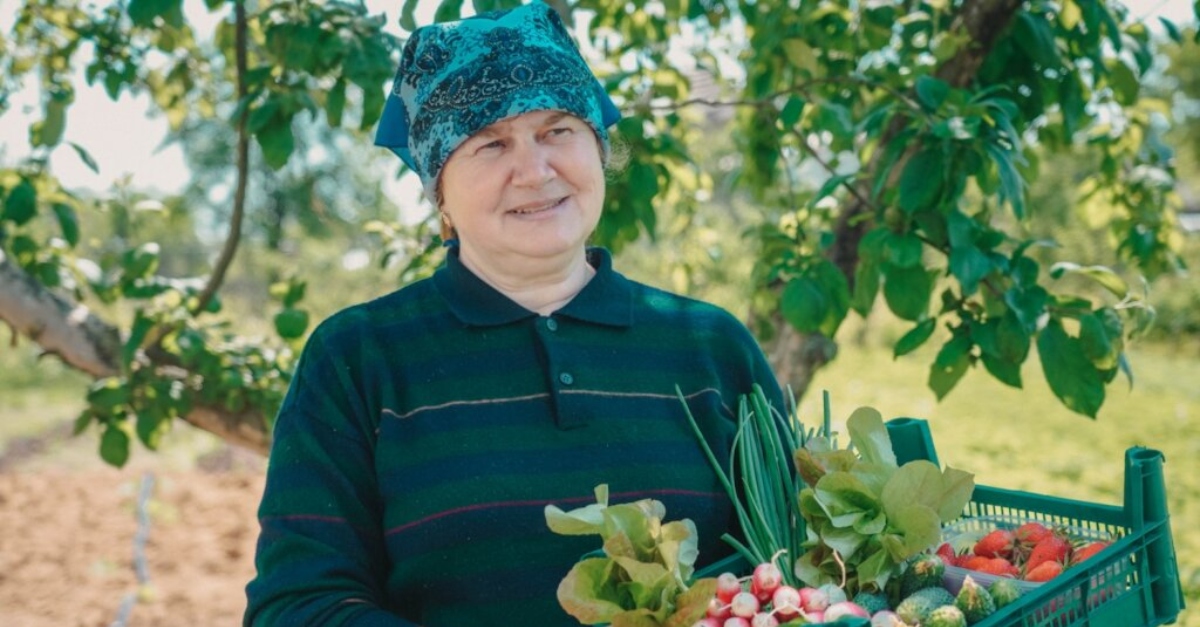一位身穿绿蓝相间衬衫、围着蓝色图案头巾的妇女站在那里，手里拿着一箱蔬菜。背景是一棵树