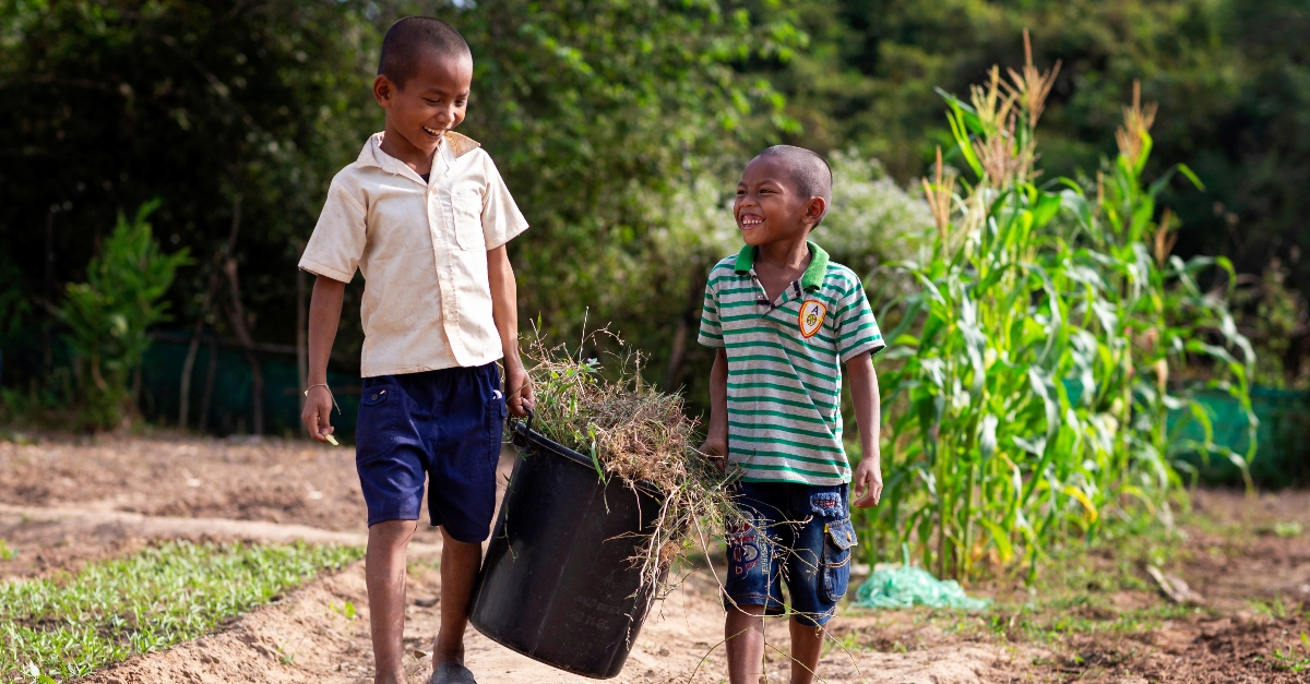 两个小孩微笑着，一边拿着一个装满草和杂草的黑桶。社区领导