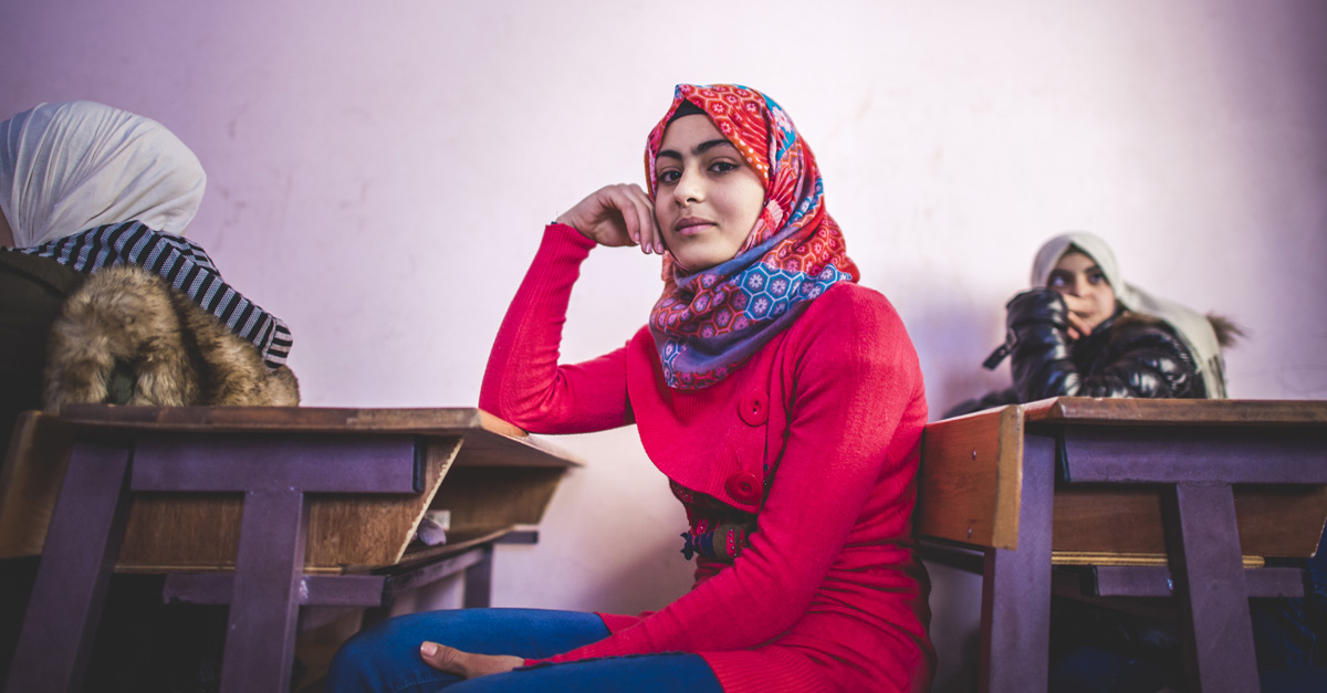 一位身穿粉色衬衫的年轻女子坐在学校的课桌前。叙利亚难民危机真相