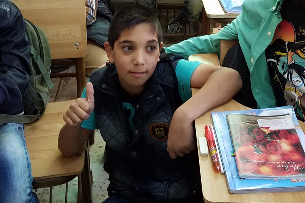 一个穿着黑色背心的小男孩坐在课桌前，竖起大拇指。其他学生在他周围的课桌上