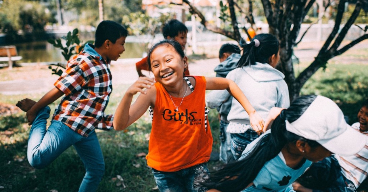 一个穿着橙色背心的女孩微笑着和其他孩子玩捉人游戏。