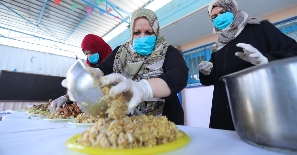 戴着蓝色面具和绿色图案头巾的纳贾夫人在一个地方端上米饭。两名戴着蓝色口罩的志愿者厨师站在她旁边。