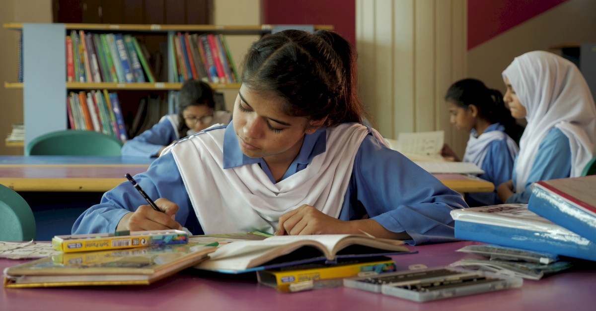 一个穿着蓝白相间制服的学生在课桌上画画时低头看着。其他女学生坐在她后面看书。