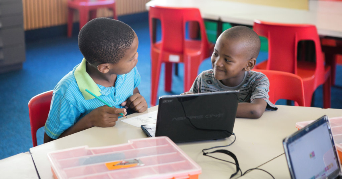 两个小男孩坐在课桌前，身后是一台笔记本电脑。背景是红椅子和一张桌子。