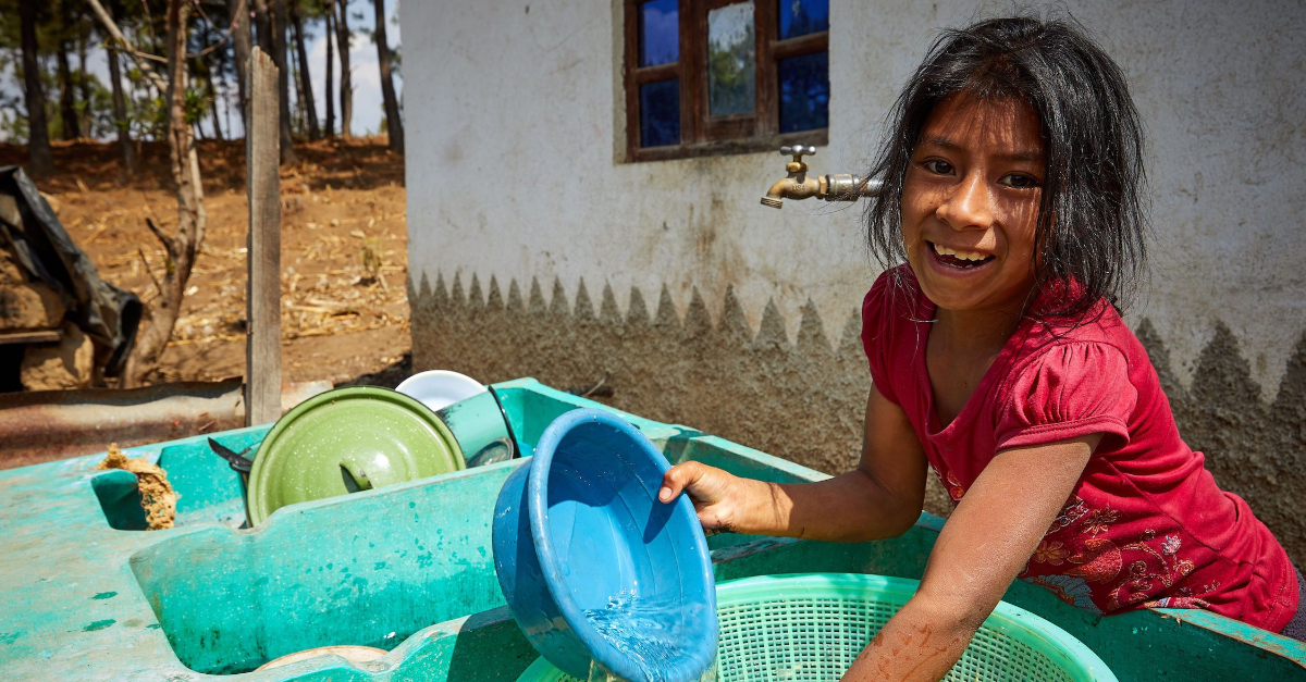 一个小女孩站在水池前，手里拿着一只蓝色的碗。背景是一座房子和一个水龙头。