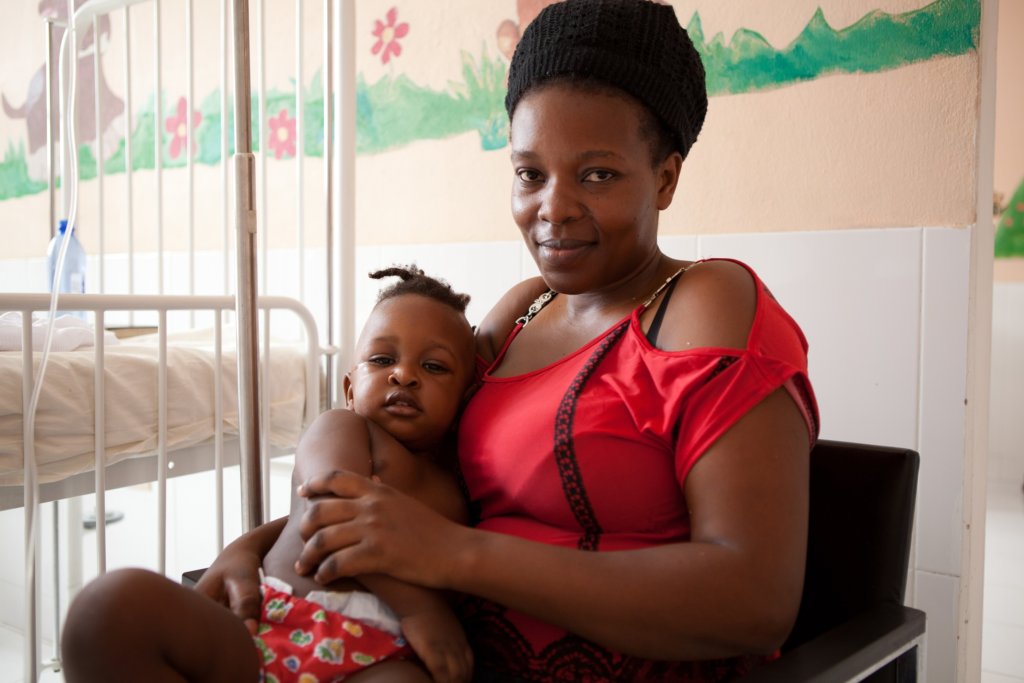 一位身穿红色衬衫的母亲将婴儿抱在膝上，旁边是医院的病床。他们身后的墙上画着花和绿草。