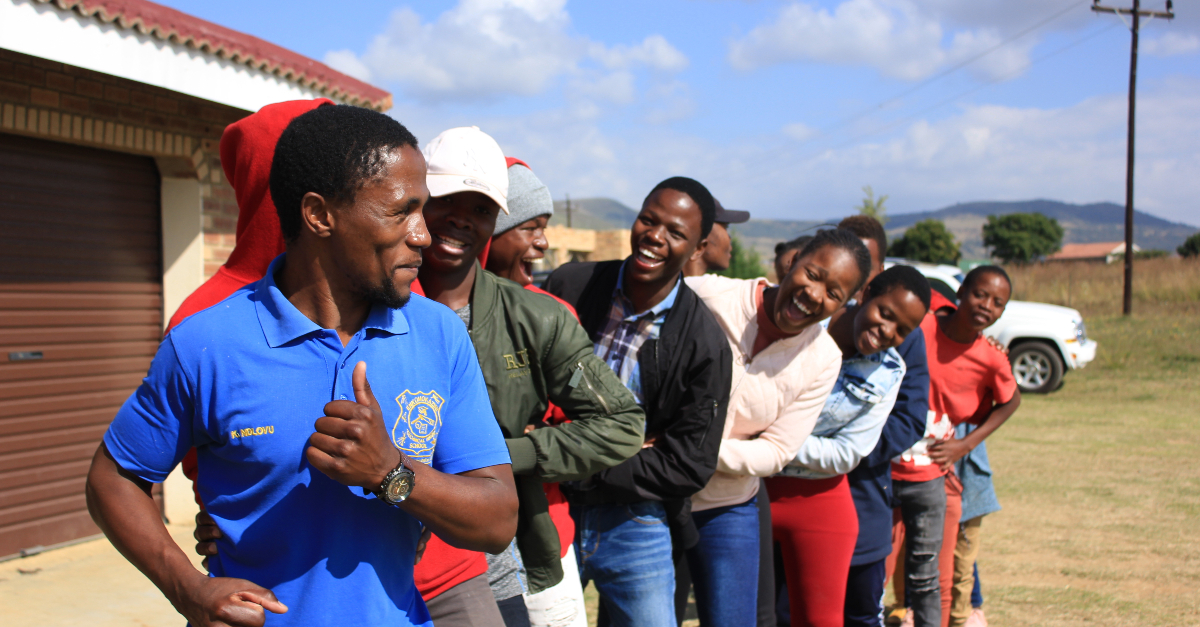 laughing people in a line following the smiling leader, house and mountainous fields in the background. Engage employees GivingTuesday