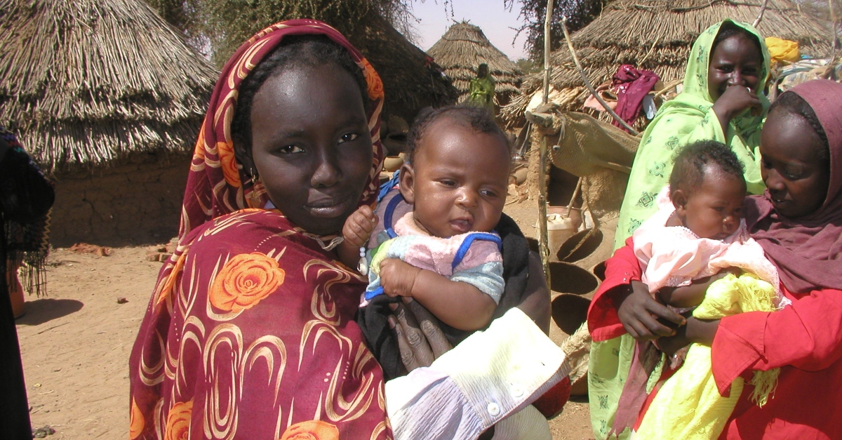在苏丹达尔富尔地区，一名妇女抱着一个婴儿。背景是其他带着孩子的妇女和泥房。